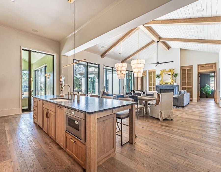 Spacious open-concept kitchen and living area with vaulted wood-beam ceilings, natural wood flooring, modern island with stone countertop, and elegant lighting, designed by Group 3 Designs.